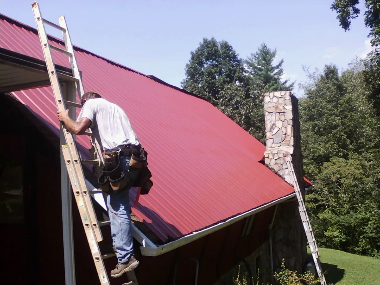 Roofer on a ladder beside a red metal roof and stone chimney — Soffit & Fascia Repair by Roofing Brothers in Ellicottville